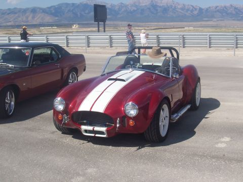 1973 Ford Mustang - A red convertible with dual white stripes sits on pavement, with another car and mountains behind it.