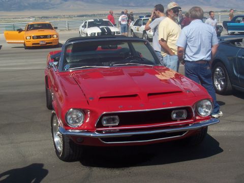 1973 Ford Mustang - A red convertible with hood scoops is parked at an outdoor car show with other vehicles.