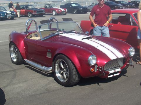 1973 Ford Mustang - A red AC Cobra with dual white racing stripes and side exhaust pipes is parked at an outdoor car show.
