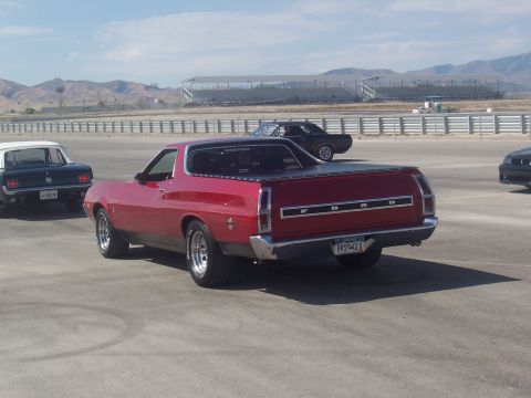 1973 Ford Mustang - A red 1970 Ford Ranchero is parked on asphalt with other vehicles and grandstands in the background.
