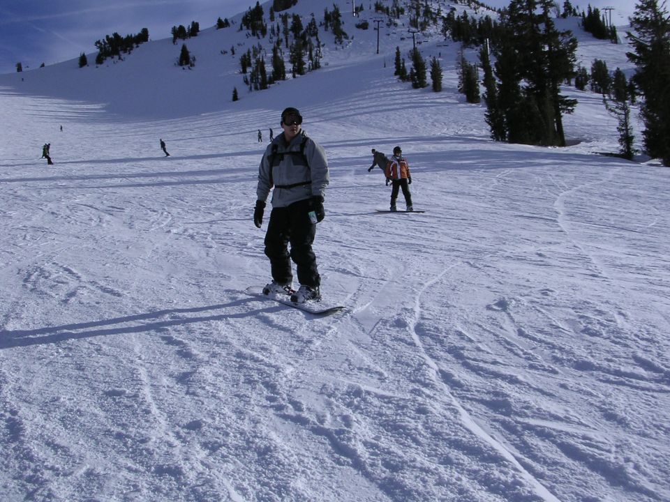 Person snowboarding down a snowy mountain slope with trees and ski lifts in the background