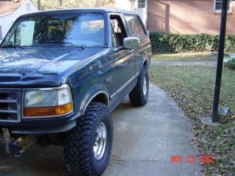 1995 Ford Bronco - Three-quarter front view of a dark green Ford Bronco on a driveway wearing new aggressive mud-terrain tires on polished five-spoke wheels,…