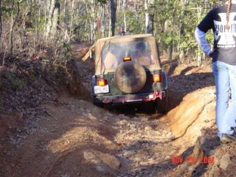 1995 Ford Bronco - Rear view of a khaki-green Jeep Wrangler Sahara with a tan soft top working through a deep eroded rut on a red-clay forest trail. The tailg…
