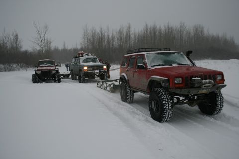 2003 Jeep TJ - A red Jeep Cherokee XJ, modified for off-road use, tows a sled through a snowy landscape with other vehicles present.
