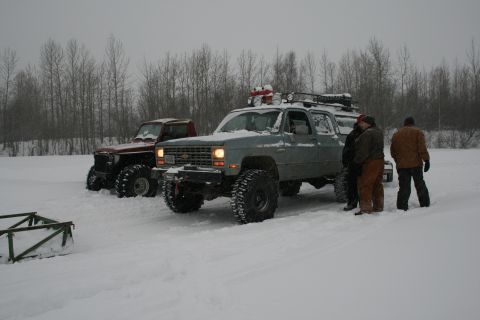 2003 Jeep TJ - Two heavily modified trucks are parked in a snowy field with several people observing.