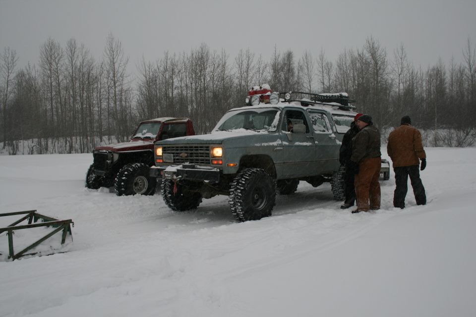 Two lifted trucks, one gray SUV and one red pickup, parked in snow with people standing nearby
