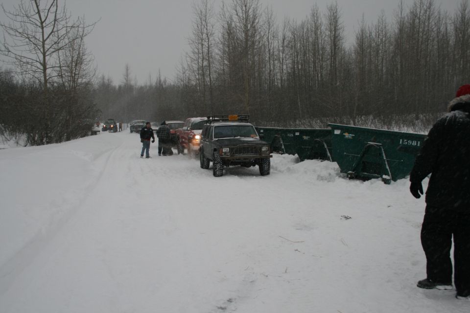 Several SUVs and trucks lined up on a snowy road next to large green dumpsters. One Jeep Cherokee XJ is in the foreground