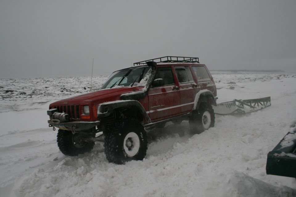 Red Jeep Cherokee XJ with snorkel and roof rack driving in snow, pulling a trailer