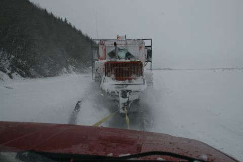 2003 Jeep TJ - Snow plow truck being towed on a snowy road, with trees on the left and a vast snowy expanse ahead.
