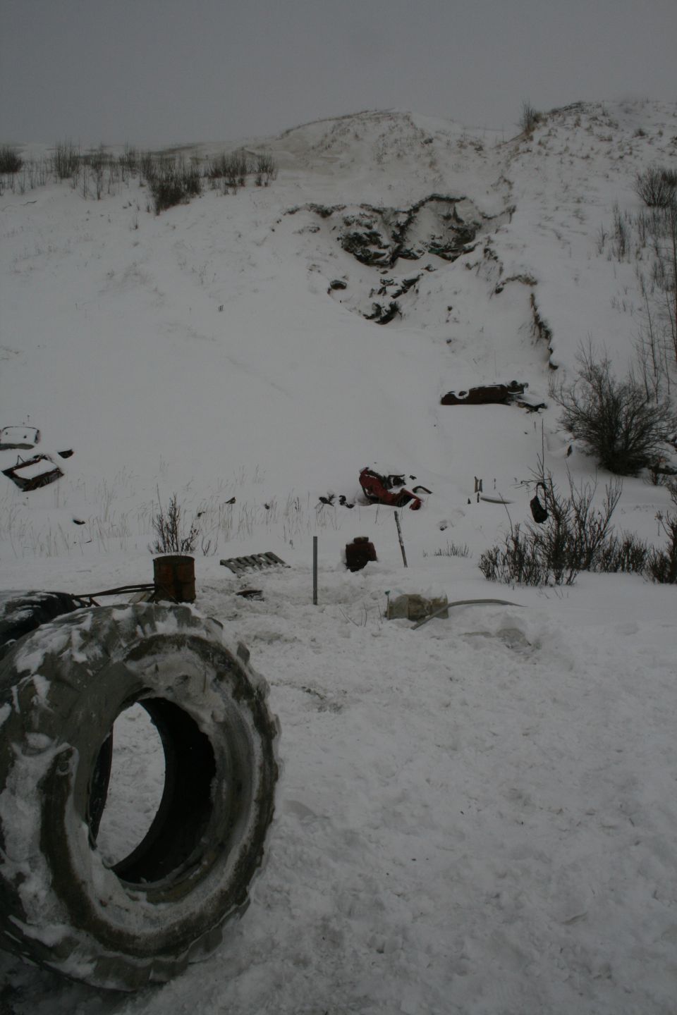 Large tire in foreground, snowy hillside with scattered debris like red metal and barrels in background