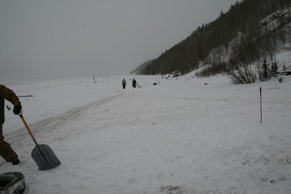 Snowy landscape with two people walking in the distance and a person shoveling in the foreground
