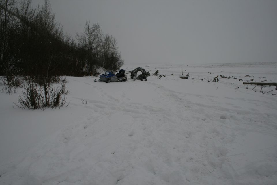 Snowmobile parked in snow next to large tires and debris on a cloudy day