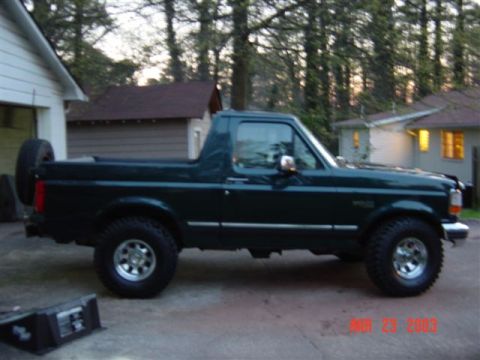 1995 Ford Bronco - A dark green full-size Ford Bronco with its top removed, side profile in a driveway at dusk, lifted on chrome wheels with a spare tire moun…