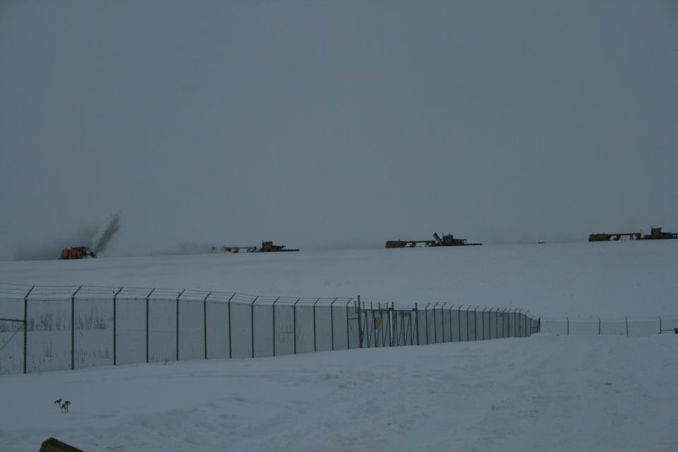 Several yellow snow blowers and other heavy equipment operate on a vast, snow-covered field under a cloudy sky, with a chain-link fence in the foreground