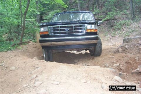 1995 Ford Bronco - Head-on view of a dark green Ford Bronco straddling a washed-out rut on a red-dirt forest trail, the front axle flexed with one tire droppe…