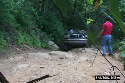 1995 Ford Bronco - Dark green Ford Bronco picking its way down a rocky, off-camber trail in dense green woods, shot head-on through overhanging branches from…