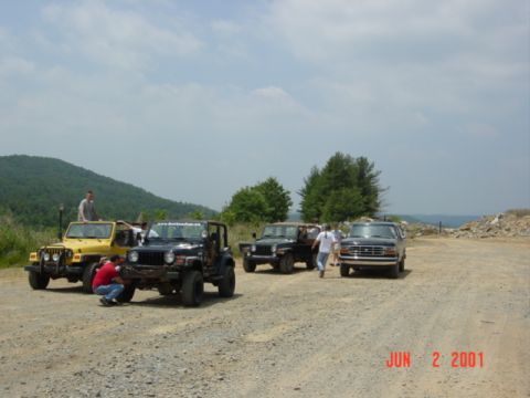 1995 Ford Bronco - Four 4x4s staged on a wide gravel road above a green mountain valley: a yellow Jeep Wrangler, a black Wrangler with a windshield banner, an…