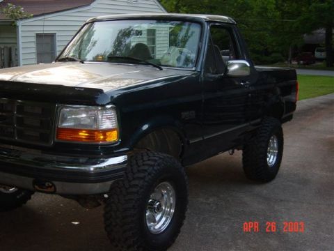 1995 Ford Bronco - Dark green Ford Bronco with the hardtop removed behind the cab, shown from the driver-side front three-quarter angle in a driveway. It sits…