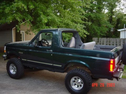 1995 Ford Bronco - A dark green Ford Bronco full-size SUV with its removable hardtop off, shown in side profile with a suspension lift, oversized mud-terrain…