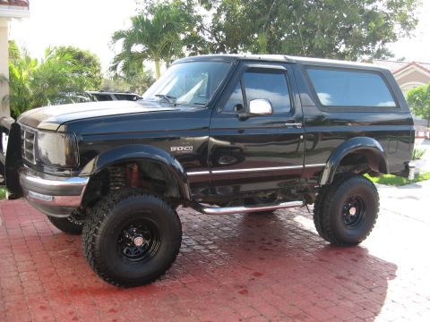 1994 Ford Bronco - This lifted black Ford Bronco is parked on a red brick surface with palm trees in the background.