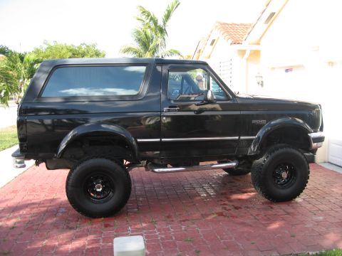 1994 Ford Bronco - This black Ford Bronco SUV is shown from the side, parked on a red brick driveway with palm trees in the background.