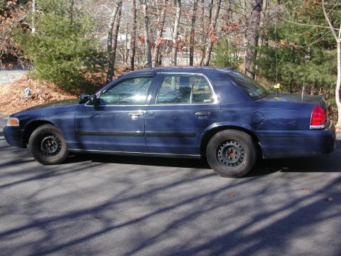 1999 Ford Crown Victoria - Dark blue sedan parked on a paved surface with trees and shadows visible.