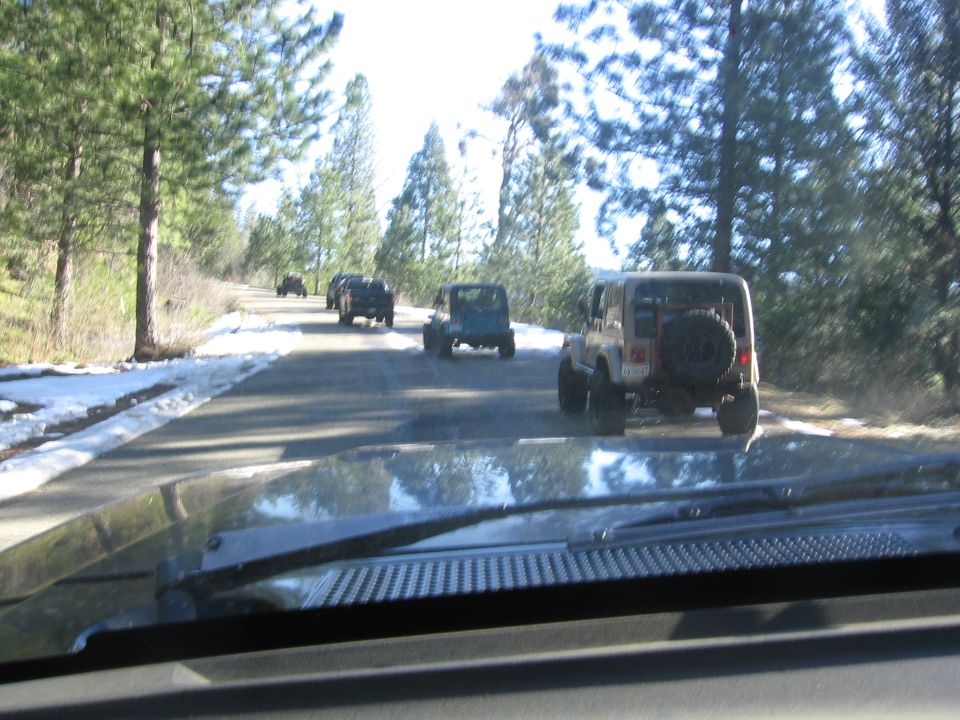 Tan Jeep Wrangler with spare tire on rear, driving on a paved road with snow patches