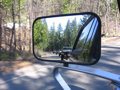 1989 Ford Bronco - A convoy of SUVs travels down a road lined with trees, as seen reflected in a side mirror.