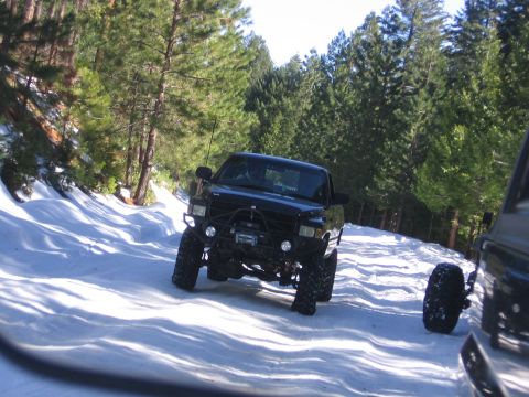 1989 Ford Bronco - A black off-road truck navigates a snow-covered path through a forest, with another vehicle visible to the right.