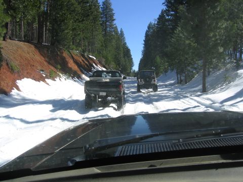 1989 Ford Bronco - Two vehicles navigate a snowy path surrounded by pine trees under a clear blue sky.
