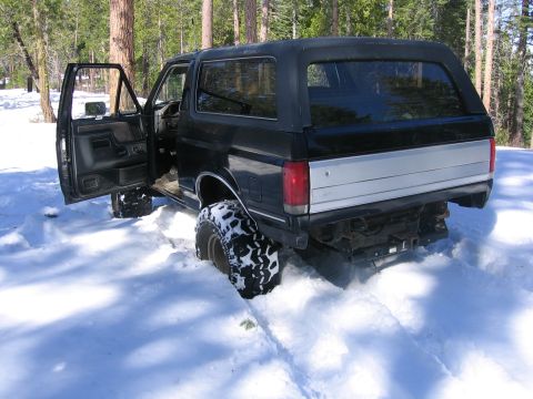 1989 Ford Bronco - Black Ford Bronco stuck in snow with the driver's door open, showing off its off-road tires.