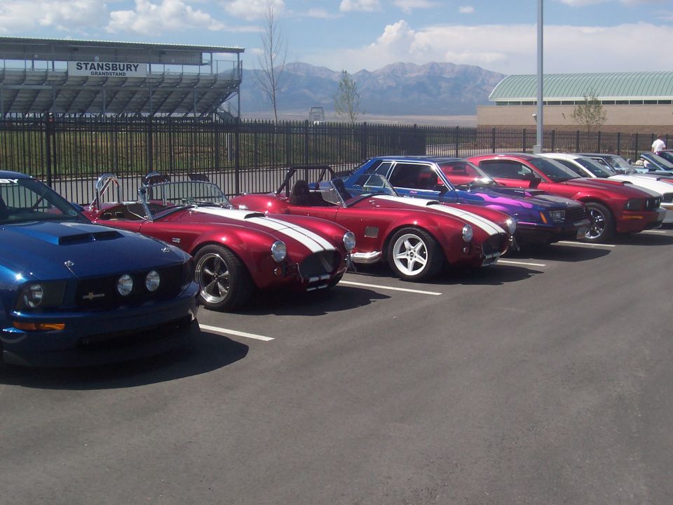 Several sports cars, including a blue Mustang and red Cobras, parked in a lot with a grandstand in the background