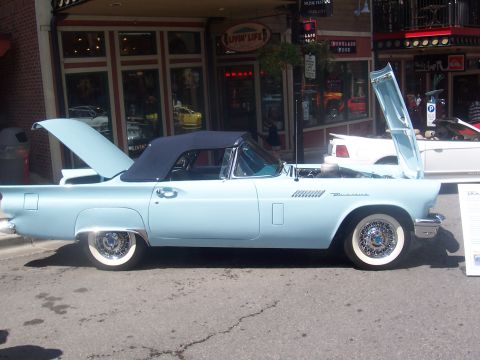 1973 Ford Mustang - A classic light blue convertible with its hood and trunk open is parked on a street in front of shops.