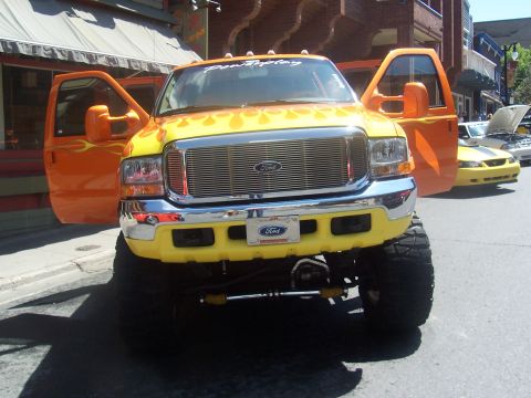 1973 Ford Mustang - A lifted Ford truck with custom flame paint and open doors is parked on a street, with another yellow car visible in the background.