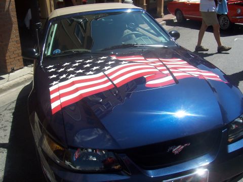 1973 Ford Mustang - A blue convertible with an American flag decal on its hood is parked on a street, with another car in the background.