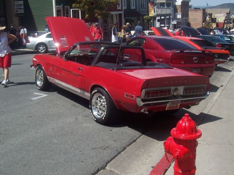 1973 Ford Mustang - A classic red convertible with Shelby badging is parked on a street with its hood open, alongside other vehicles.