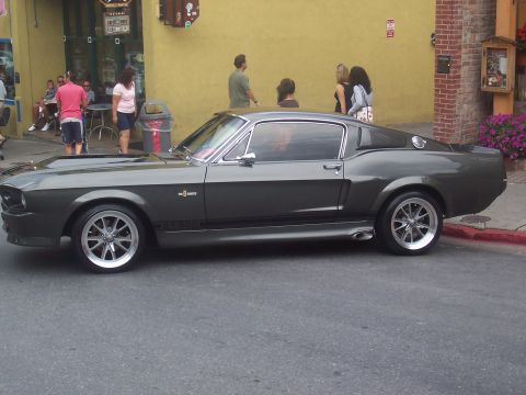 1973 Ford Mustang - A classic dark gray coupe with racing stripes is parked curbside in front of a building.