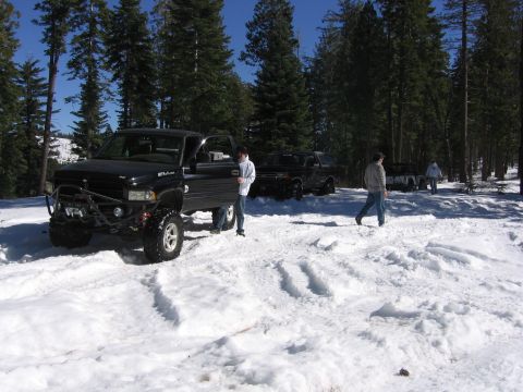 1989 Ford Bronco - Several pickup trucks are parked in a snowy wooded area with people standing around.