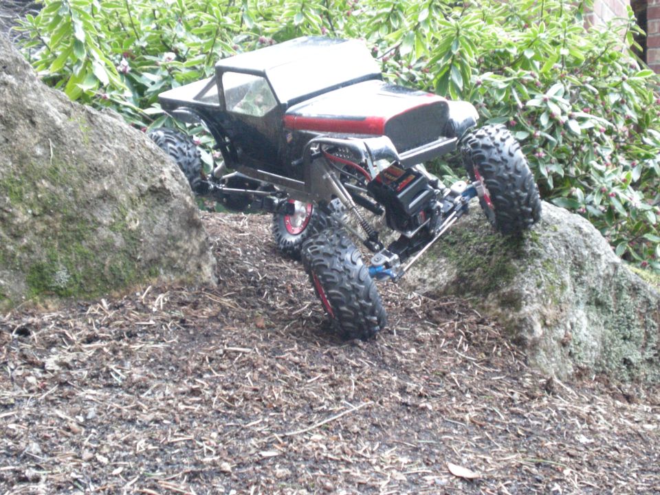 RC rock crawler climbing over rocks and dirt with green foliage in background