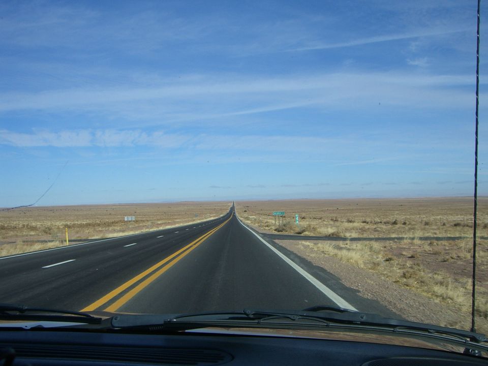 Desert highway stretching to the horizon under a blue sky with scattered clouds. Dry grass lines the roadside
