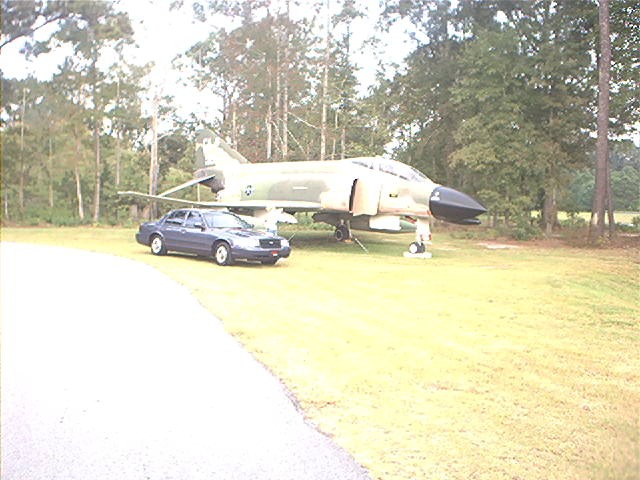 Dark blue Ford Crown Victoria parked on grass next to a camouflaged F-4 Phantom jet display