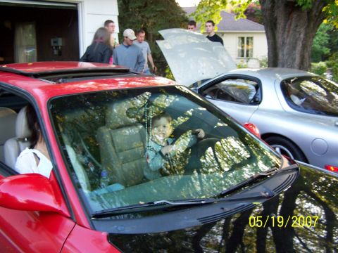 1996 Ford Probe - A baby sits in the driver's seat of a red car, with a woman in the passenger seat and other people and a silver car in the background.