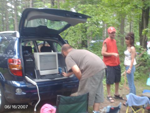 1996 Ford Probe - People gathered around a car with a computer setup in the trunk, possibly at a campsite.