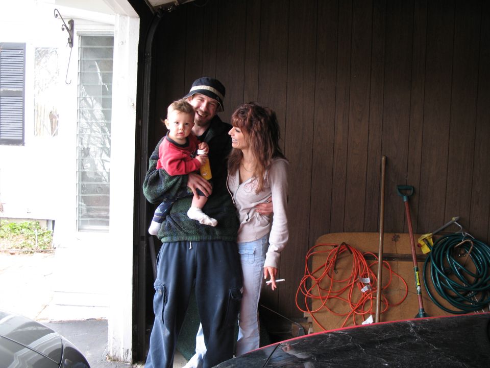 Man holding a baby, woman standing next to him, in a garage with tools and supplies
