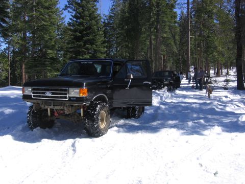 1989 Ford Bronco - A lifted black Ford Bronco sits in the snow with its door open, joined by other vehicles and people in a wooded area.