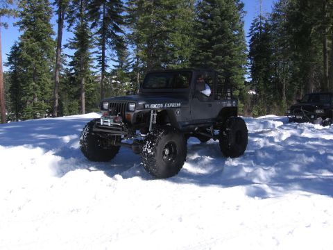 1989 Ford Bronco - A dark Jeep Wrangler, modified with "Rubicon Express" branding, navigates a snowy terrain with another vehicle visible in the background.