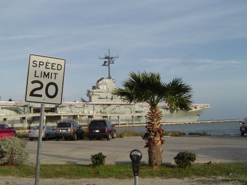1989 Ford Bronco - A speed limit sign indicates 20 mph in a parking lot with several cars, a palm tree, and a large aircraft carrier in the background.