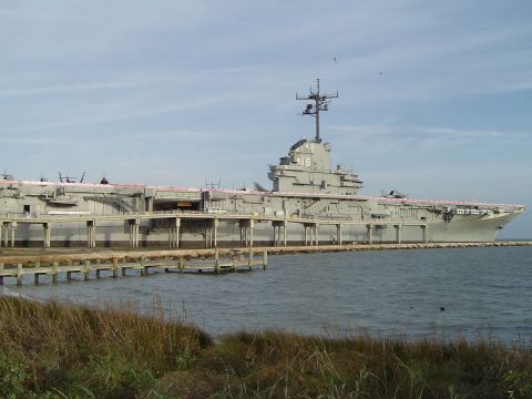 1989 Ford Bronco - An aircraft carrier is docked at a pier, with a wooden walkway and grassy shore in the foreground.