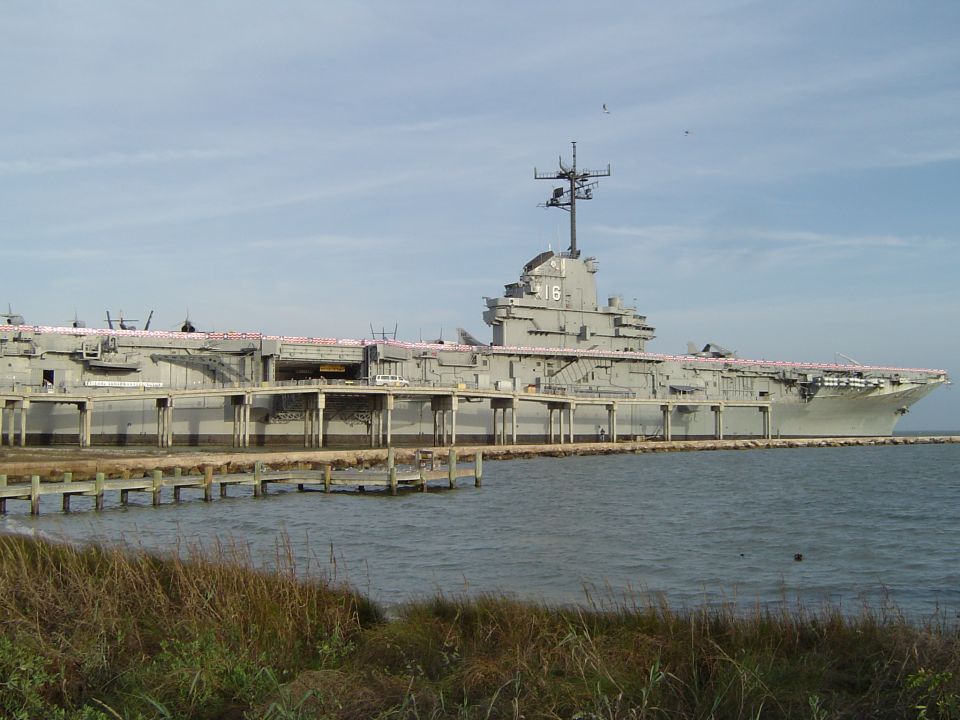 Aircraft carrier docked at a pier, with a wooden walkway in the foreground and grass. The number 16 is visible on the ship