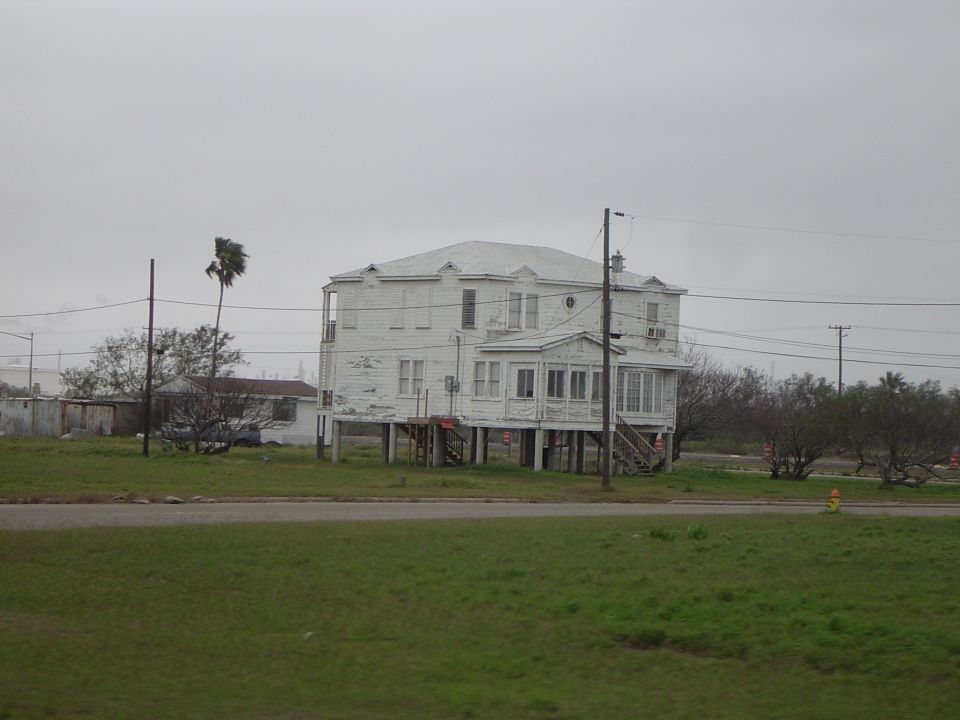 White house on stilts with peeling paint, seen from across a grassy field under an overcast sky